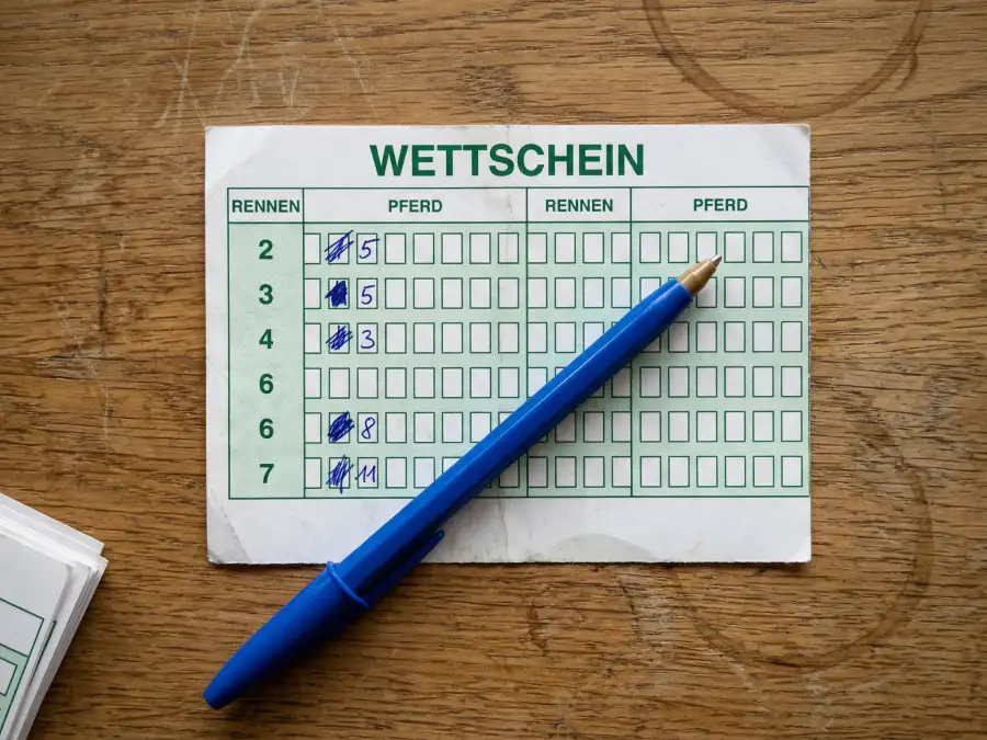 Ausgefüllter Wettschein für eine Pferdewette mit angekreuzten Feldern und Kugelschreiber auf einem Tresen der Rennbahn